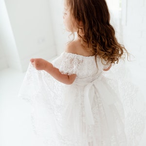May include: A young girl wearing a white off-the-shoulder lace dress with a satin ribbon tied at the waist. The dress has a flowing skirt and is perfect for a special occasion.