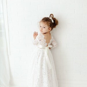 May include: A white lace dress featuring long sleeves, a deep scoop back, and a satin ribbon tied in a bow at the waist. The floor-length skirt is made of intricate floral lace, and the bodice has a scalloped neckline and back edge detail.