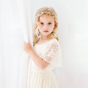 以下が含まれることがあります： 白いレースのドレスを着て、金色の花の冠をつけた若い女の子。彼女は白いカーテンの横に立ってカメラを見ている。