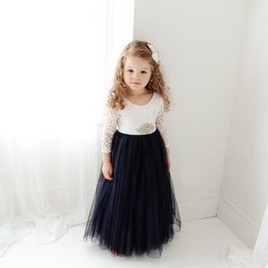 May include: A young girl wearing a white lace top with a navy blue tulle skirt and a sparkly flower embellishment on the waist.