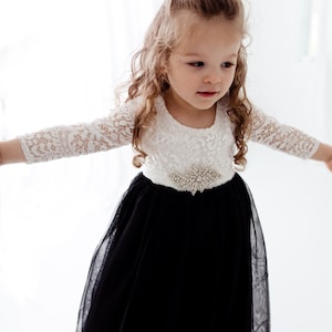 May include: A dress featuring a white lace bodice with three-quarter length sleeves and a full black tulle skirt. The waist is accented with a decorative silver-toned brooch made of clear crystals. A small white pearl-adorned hair accessory is visible.