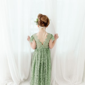 K&ouml;nnte beinhalten: Ein junges M&auml;dchen tr&auml;gt ein salbeigr&uuml;nes Spitzenkleid mit tiefem R&uuml;cken und einer Blumenkrone. Sie steht vor einem wei&szlig;en Vorhang und schaut aus einem Fenster.