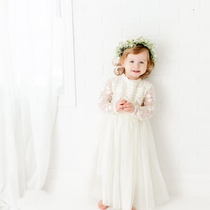 May include: A floor-length off-white dress with long sheer lace sleeves, a high neckline with scalloped trim, and a layered tulle skirt. The garment is paired with a floral crown featuring small white blossoms and green foliage accents.