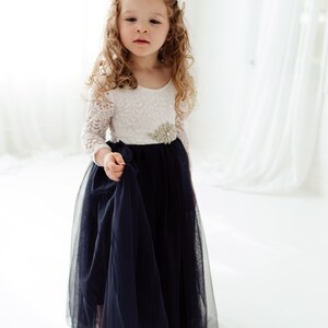 May include: A young girl wearing a white lace long-sleeved dress with a navy blue tulle skirt and a silver embellished belt.
