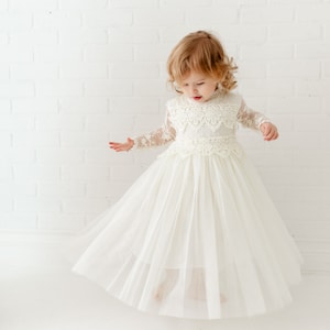 May include: A cream-colored dress featuring long, sheer lace sleeves with floral patterns, a high neckline, and a bodice adorned with intricate scalloped lace trim. The skirt is constructed from multiple layers of soft, flowing tulle fabric.