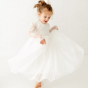 May include: A young girl wearing a white tulle dress with lace detailing and long sleeves. She is twirling and smiling.