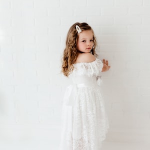 May include: A young girl wearing a white off-the-shoulder lace dress with a satin sash. She has curly brown hair and is wearing a pearl hair clip.