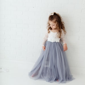 May include: A young girl wearing a white lace top with a tulle skirt in a light blue colour. The skirt is long and flowing, and the girl is standing in front of a white brick wall.