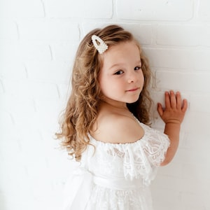 May include: A young girl with curly blonde hair wearing a white off-the-shoulder lace dress with a satin sash. She is wearing a pearl hair clip and has a sweet expression on her face.