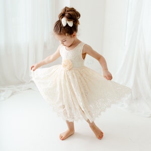 May include: A young girl wearing a white lace dress with a flower accent on the waist. She is twirling in a white room with white curtains.