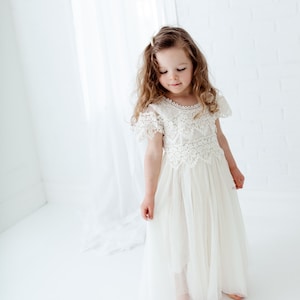 May include: A young girl wearing a cream-colored lace dress with a tulle skirt. The dress features a detailed lace bodice and short sleeves. The girl has long, wavy brown hair and is standing in a bright, white room.