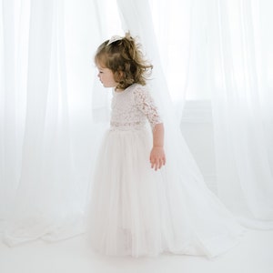 May include: A young girl in a white lace dress with a tulle skirt stands in front of a white curtain.