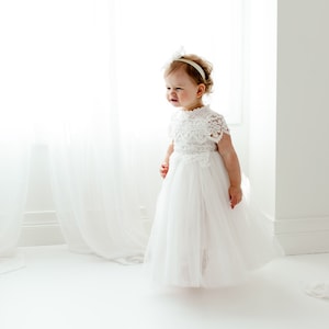 May include: A white lace and tulle dress with a matching headband. The dress has a detailed lace bodice and a flowing tulle skirt. The child is smiling, standing in front of a bright window.