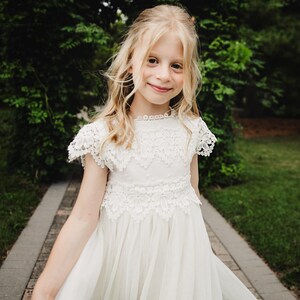 May include: A young girl with blonde hair smiles, wearing a white dress with lace detailing. The dress features a high neckline and short sleeves. She stands on a stone path, with green foliage in the background.