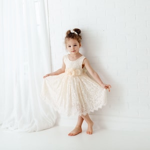 May include: A young girl wearing a white lace dress with a peach-colored flower accent at the waist. She is standing in front of a white brick wall.