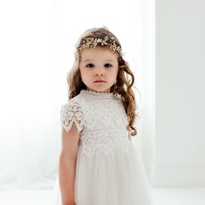 May include: A young girl in a white lace dress and a gold floral headband. The dress has a high neckline and lace detailing on the sleeves and bodice. The girl has long, curly brown hair.