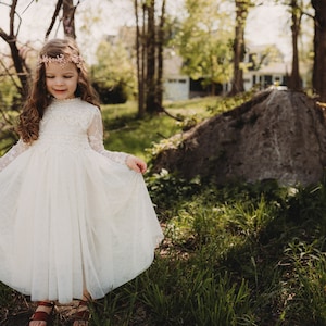 K&ouml;nnte beinhalten: Ein junges M&auml;dchen in einem wei&szlig;en Spitzenkleid und einer Blumenkrone steht in einem grasbewachsenen Bereich mit einem gro&szlig;en Felsen und B&auml;umen im Hintergrund.