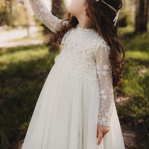 K&ouml;nnte beinhalten: Ein junges M&auml;dchen tr&auml;gt ein wei&szlig;es Spitzenkleid mit langen &Auml;rmeln. Das Kleid hat ein zartes Blumenmuster und einen vollen Rock. Das M&auml;dchen steht in einem grasbewachsenen Bereich mit B&auml;umen im Hintergrund.