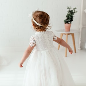 May include: Two young girls in white dresses. One girl is in the foreground, wearing a dress with lace sleeves and a tulle skirt. The other girl is seated, wearing a similar dress and a floral crown. The background is a white room with a small table and a plant.