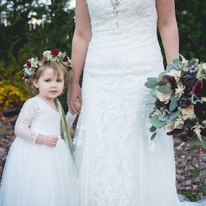 May include: A young girl in a white dress and flower crown holds hands with a bride in a white lace wedding dress. The bride is holding a bouquet of burgundy, white, and green flowers.