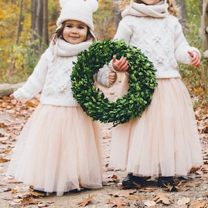 May include: Two young girls wearing white knit sweaters and matching tulle skirts with white pom-pom hats. The girls are holding a green boxwood wreath.