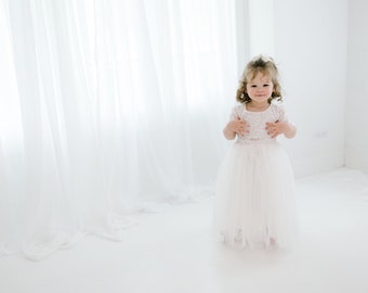 Op de afbeelding: Een peuterjurk met een wit kanten lijfje met korte mouwen en een ronde halslijn, gecombineerd met een lange, gelaagde tule rok in een bijpassende witte kleur. Het kledingstuk is voorzien van een delicaat bloemmotief op het kanten lijfje.