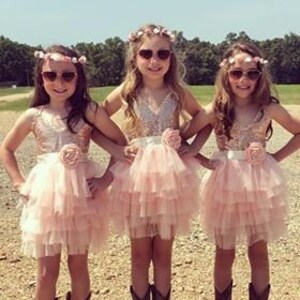 以下が含まれることがあります： 花冠とサングラスをつけた3人の女の子が、ピンクと白のキラキラしたチュールスカートのドレス、花のサッシュ、カウボーイブーツを履いています。