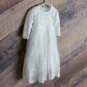 May include: White lace baby dress with long sleeves and a round neckline. The dress is hanging on a white hanger against a wooden background.