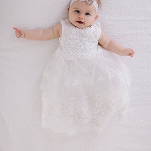May include: A baby girl wearing a white lace dress and a headband with a rhinestone embellishment. The baby is lying on a white blanket.