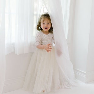 May include: A young child wearing a white lace bodice dress with a tulle skirt, smiling and peeking out from behind a sheer white curtain. The room is brightly lit with natural light.