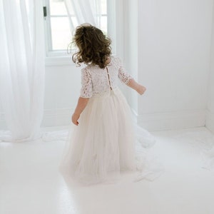 May include: A young child wearing a white lace dress with a tulle skirt, standing near a window with sheer white curtains. The dress has short sleeves and a detailed lace bodice.