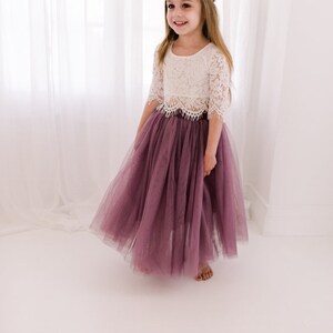 May include: A two-piece outfit consisting of a white lace top with three-quarter length sleeves and a scalloped hem, paired with a floor-length, deep mauve tulle skirt. A matching deep mauve velvet bow headband is worn on the head.