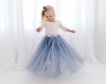 Op de afbeelding: Een tweedelige outfit bestaande uit een wit kanten top met lange mouwen en een geschulpte zoom en een volumineuze, vloerlange tule rok in stofblauw. De top heeft ingewikkelde bloemmotieven en de rok bestaat uit meerdere lagen zachte stof.