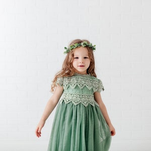 May include: A young girl wearing a sage green tulle dress with lace detailing and a green leaf crown. The dress has a round neckline and short sleeves.