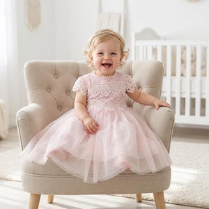 May include: A light pink dress featuring a lace bodice with short sleeves and a layered tulle skirt. The dress is worn with a thin, gold-toned headband adorned with small pearls, creating a soft and delicate appearance for a young child.