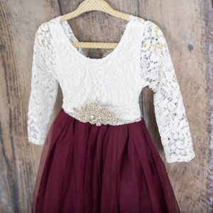 May include: A long-sleeved dress featuring a white lace bodice with a scoop neckline and a deep burgundy tulle skirt. A decorative gold-toned rhinestone embellishment is centered at the waistline where the lace bodice meets the tulle skirt.