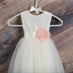 Op de afbeelding: Een witte tule jurk met een pailletten lijfje en een perzikkleurige bloem accent. De jurk heeft een satijnen band en is perfect voor een speciale gelegenheid.