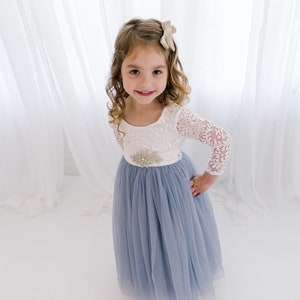 May include: A young girl wearing a white lace top with a blue tulle skirt and a sparkly embellishment at the waist. She is smiling and looking at the camera.