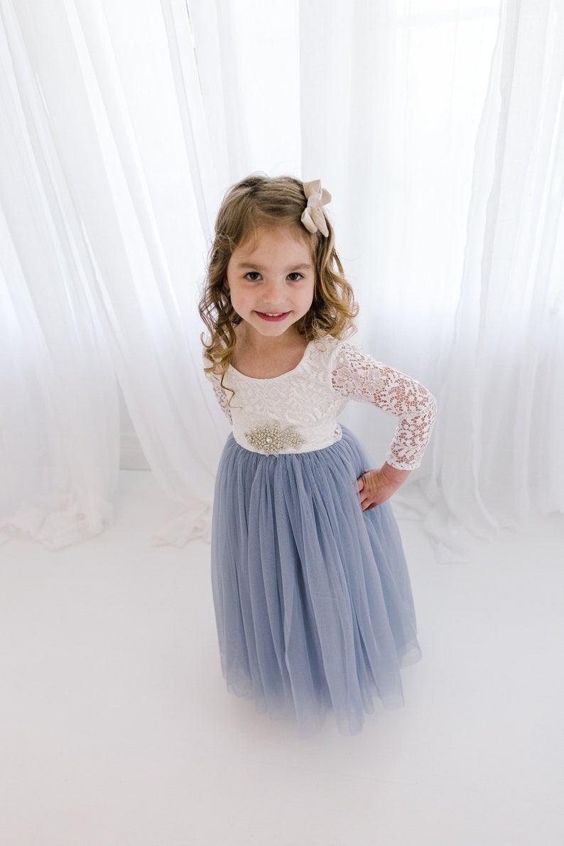 May include: A young girl wearing a white lace top with a blue tulle skirt and a sparkly embellishment at the waist. She is smiling and looking at the camera.
