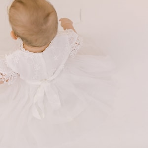 K&ouml;nnte beinhalten: Ein wei&szlig;es Babykleid mit Spitzendetails und einer Schleife an der Taille. Das Kleid ist aus einem weichen, flie&szlig;enden Stoff gefertigt und perfekt f&uuml;r einen besonderen Anlass.