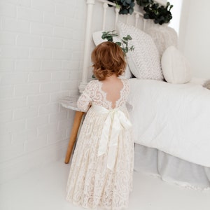 May include: A young girl wearing a white lace dress with a satin sash. The dress has a low back and long sleeves. The girl is standing in front of a white bed with a white headboard and a white bedspread.
