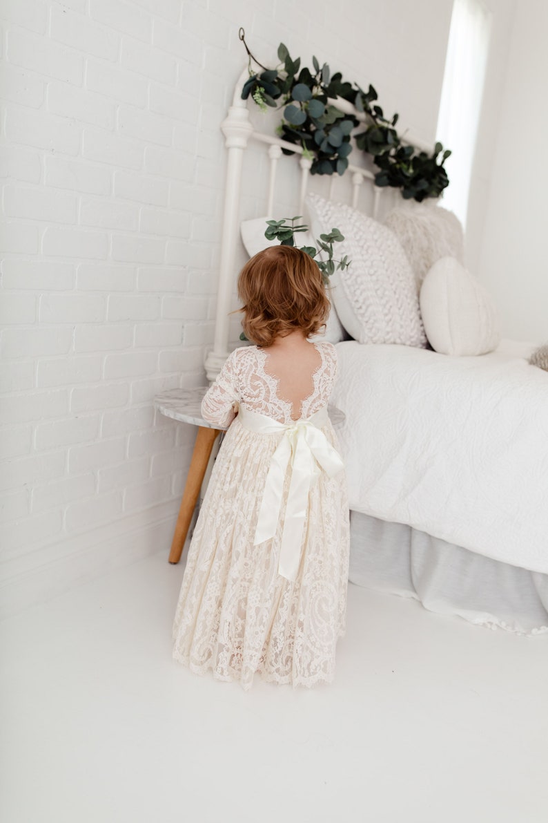May include: A young girl wearing a white lace dress with a satin sash. The dress has a low back and long sleeves. The girl is standing in front of a white bed with a white headboard and a white bedspread.