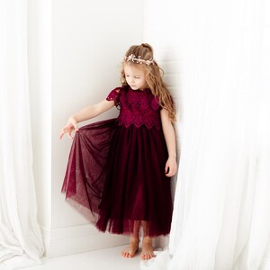 May include: A young girl wearing a burgundy tulle dress with lace detailing and a flower crown. The dress has a full skirt and short sleeves. The girl is standing in a white room with white curtains.
