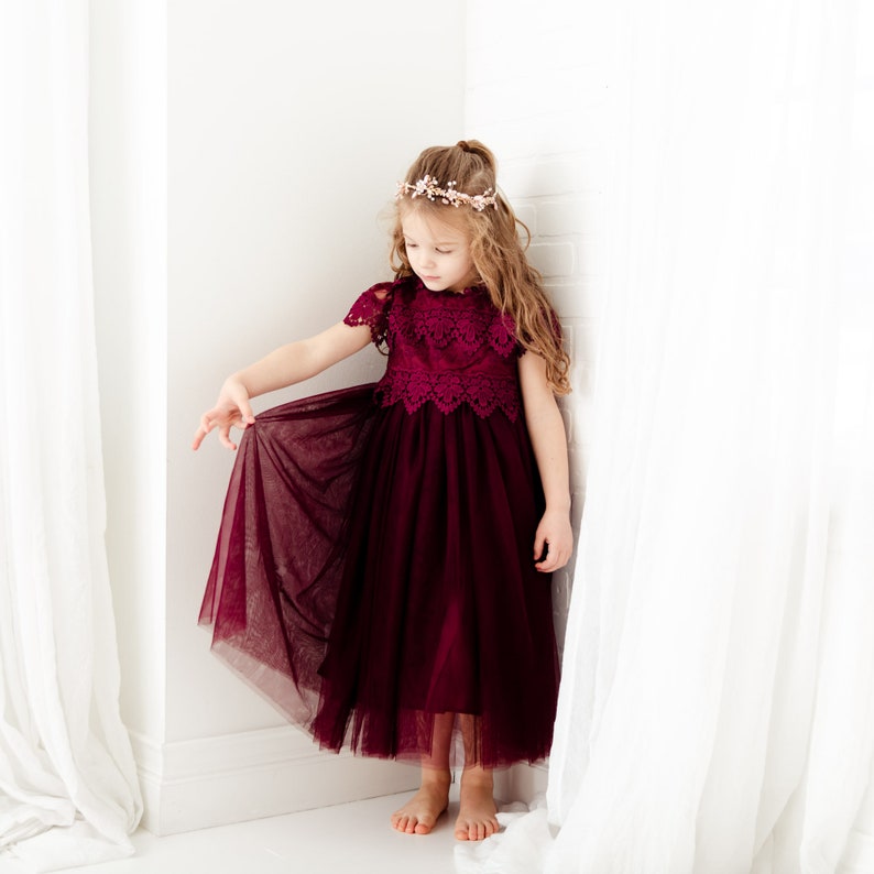 May include: A young girl wearing a burgundy tulle dress with lace detailing and a flower crown. The dress has a full skirt and short sleeves. The girl is standing in a white room with white curtains.