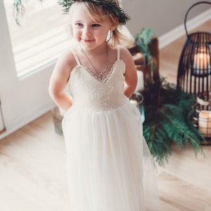 May include: A young girl wearing a white dress with a sequined bodice and a tulle skirt. She is wearing a green wreath in her hair and a silver necklace. The girl is standing in front of a window with greenery behind her.