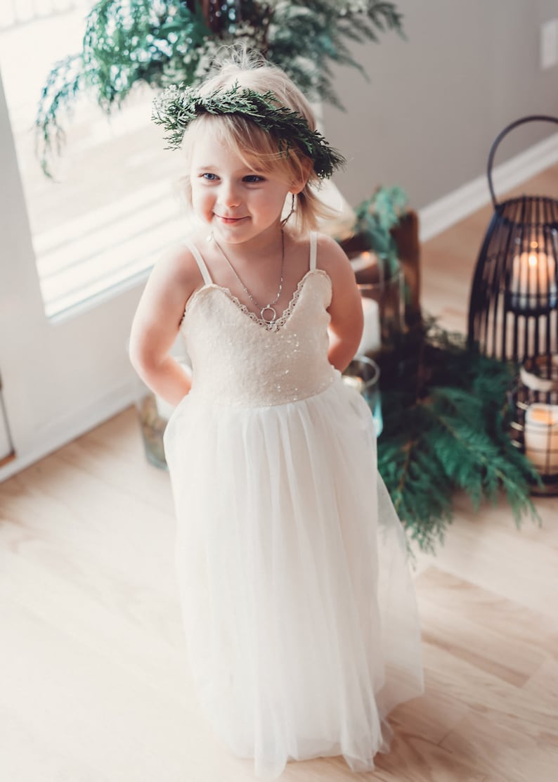 May include: A young girl wearing a white dress with a sequined bodice and a tulle skirt. She is wearing a green wreath in her hair and a silver necklace. The girl is standing in front of a window with greenery behind her.