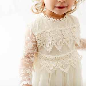 May include: An off-white dress featuring long sheer sleeves with floral embroidery, a high neckline with scalloped lace trim, and two horizontal bands of intricate floral lace appliqu&eacute; across the bodice, set against a plain white background.