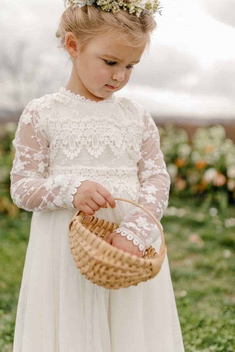 May include: A young girl in a white lace dress holds a woven basket. She is wearing a flower crown and looking down at the basket.