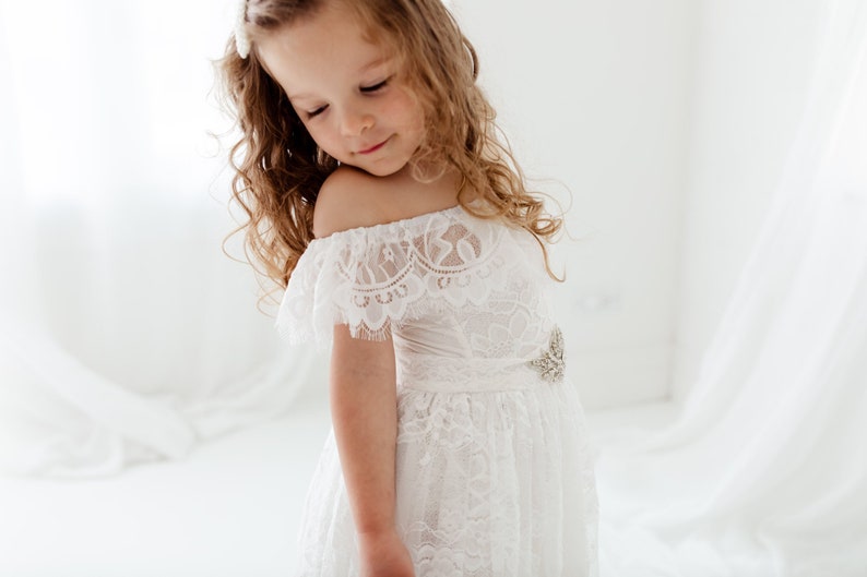 May include: A young girl wearing a white off-the-shoulder lace dress with a beaded belt. The dress has a ruffled neckline and a flowing skirt.