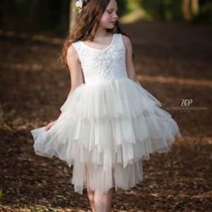 May include: A young girl wearing a white tulle dress with a floral crown. The dress has multiple layers and a high neckline. The girl is standing in a wooded area.